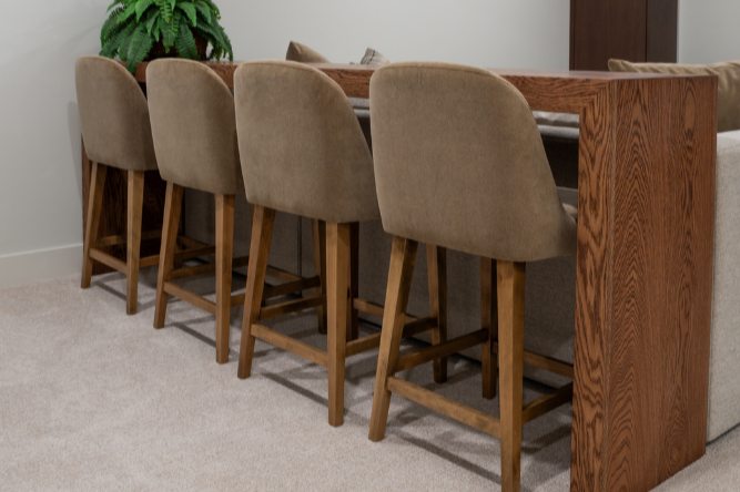 Four tan-upholstered bar stools lined up at a long wooden console table behind a sectional sofa in a finished basement.