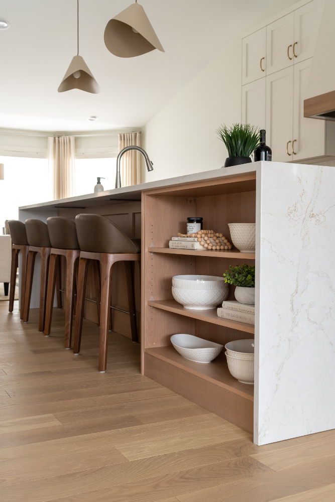A close-up view of a natural wood kitchen island with integrated open shelves holding curated ceramics, plants, and four brown leather stools.