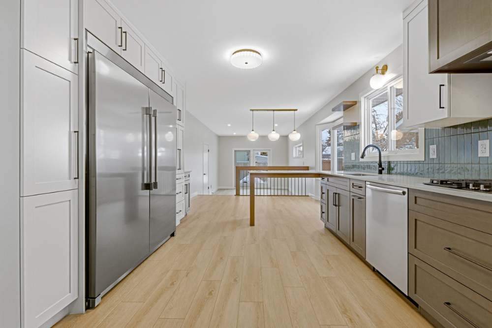 A perspective shot of a modern kitchen aisle with a large stainless steel double-door refrigerator on the left and a long counter with a black faucet and teal backsplash on the right.