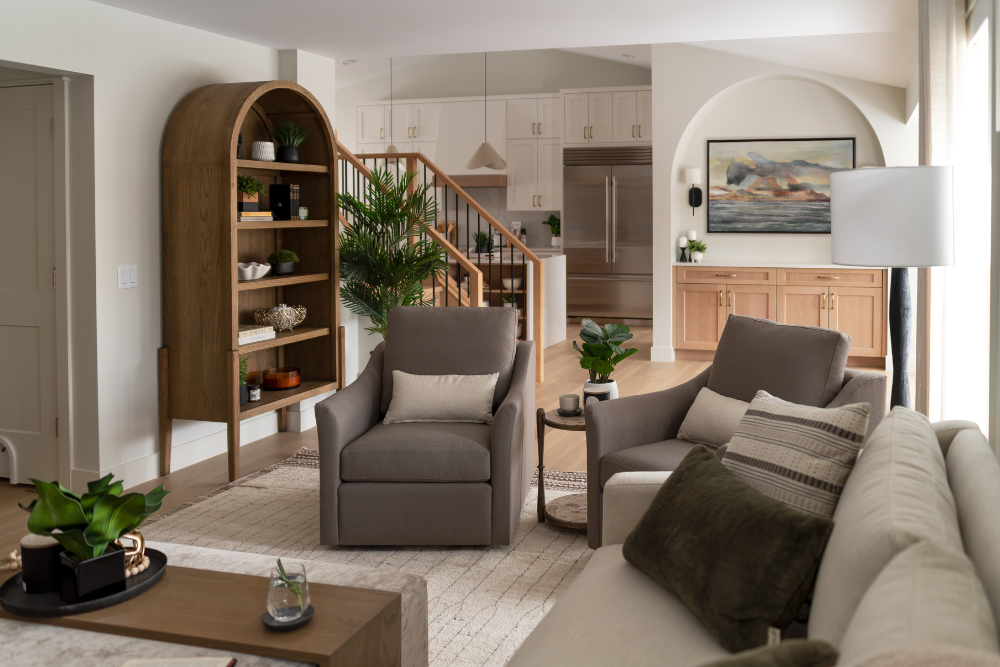 A wide view of an open-concept living space highlighting an arched wooden cabinet, grey armchairs, a sofa, and a glimpse of the kitchen.