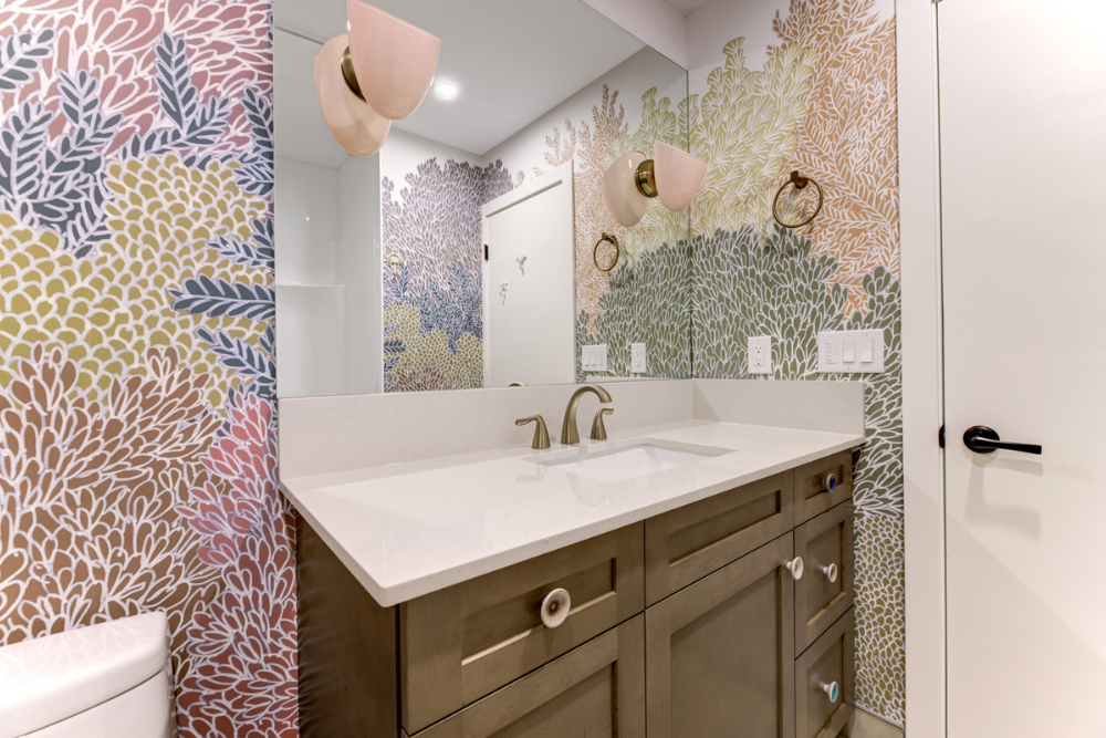 A close-up perspective of a custom light wood vanity in a bathroom with botanical wallpaper. The image focuses on the white countertop, a bronze-finish gooseneck faucet, and the varied decorative ceramic pulls on the cabinet doors and drawers. 