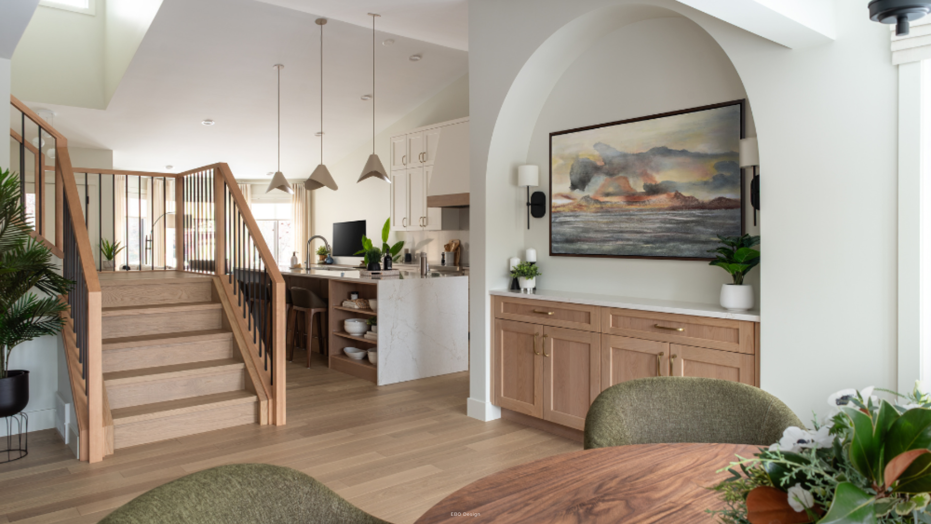 An open-concept living space featuring a modern light wood staircase with black spindles, a large arched wall niche with built-in cabinets and artwork, and a view into the kitchen with pendant lighting.