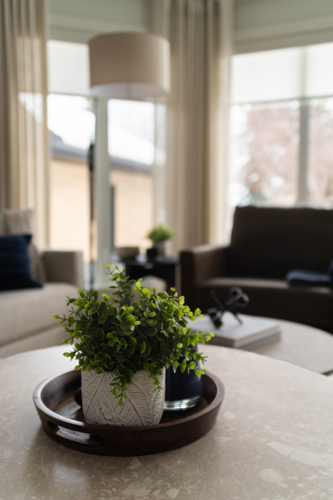 A close-up shot of a small, vibrant artificial eucalyptus plant in a textured grey pot, placed on a dark round wooden tray on a light marble coffee table in a modern living room.