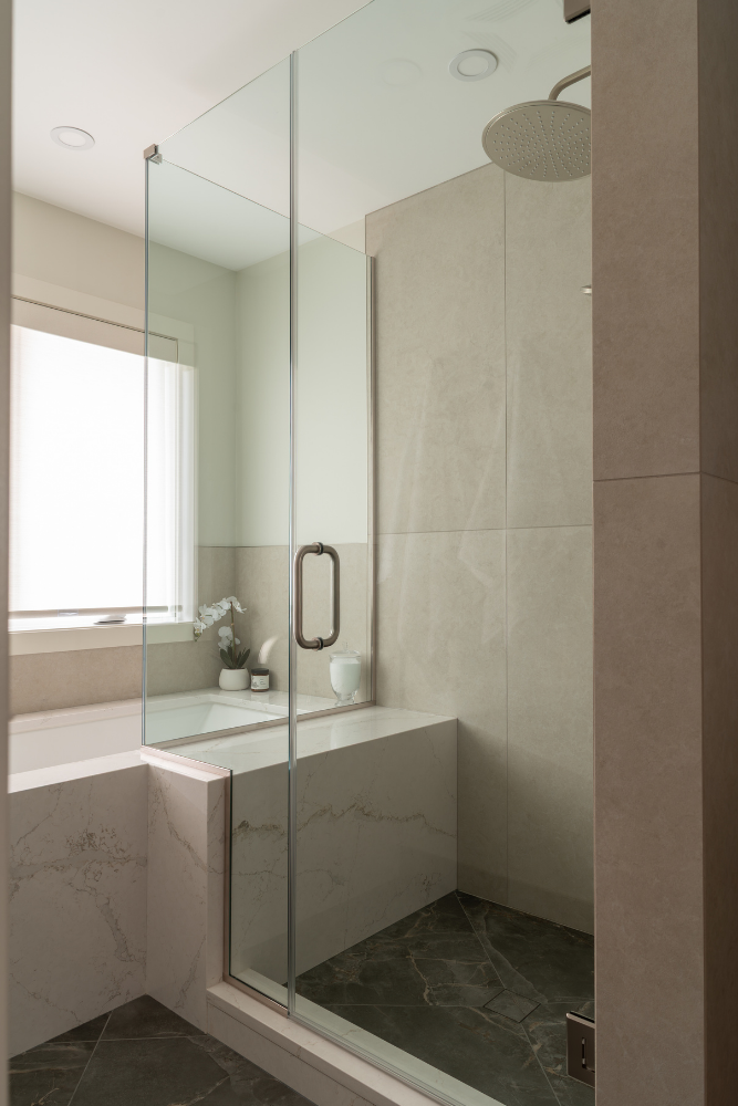 A partial view of a modern walk-in shower featuring a frameless glass enclosure with a large metal handle. Large format tiles line the walls, and a large, round rainfall showerhead is visible overhead.