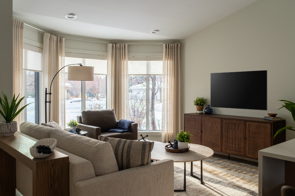 A view of an open-concept living area showing a beige sectional sofa, nesting coffee tables, a leather armchair, and a walnut media console, all near a large bay window.