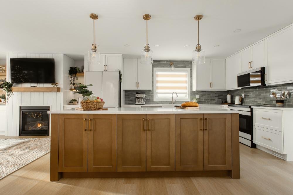 Modern open-concept kitchen renovated by EBO Design, featuring white shaker cabinets, a warm wood island, gold pendant lighting, green tile backsplash, and a cozy living room with a shiplap fireplace.