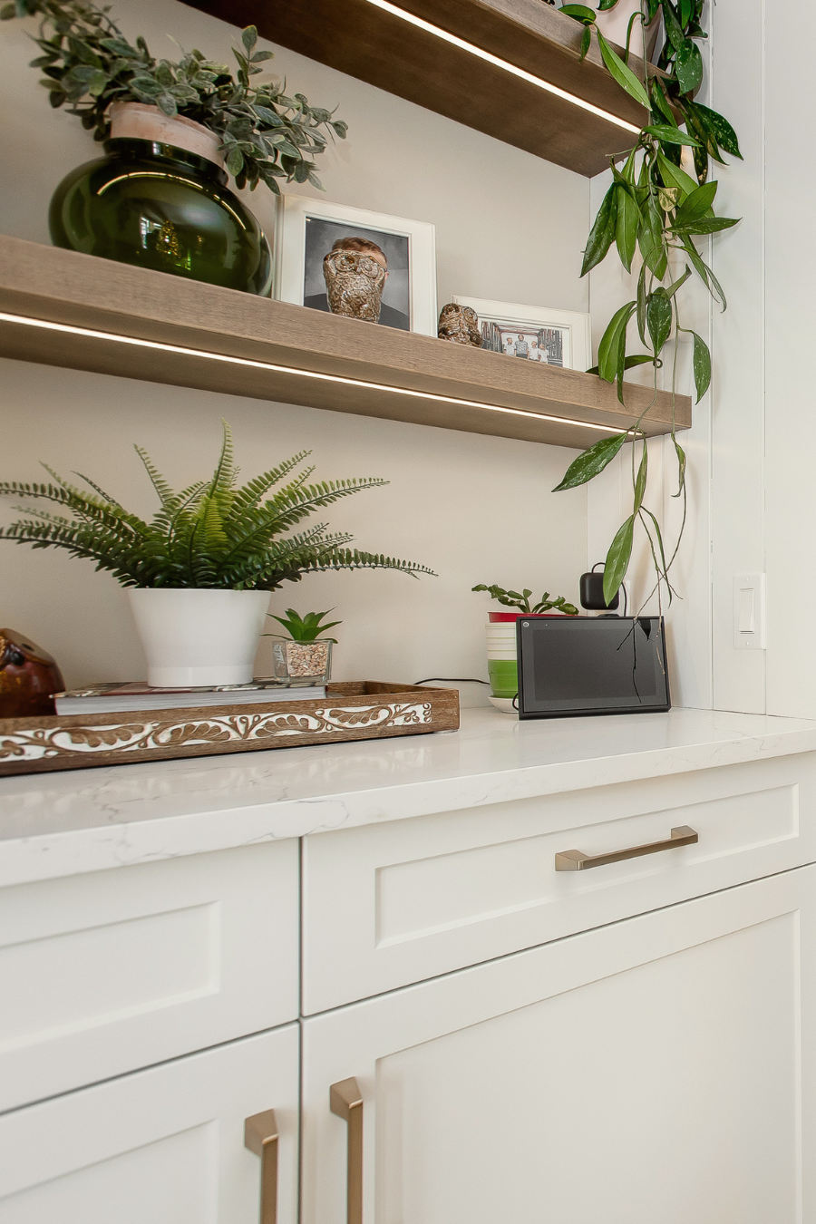 Modern living room built-in with white shaker cabinets, quartz countertops, floating wood shelves, and styled décor including greenery, framed photos, and a smart home display for a warm, personalized space.
