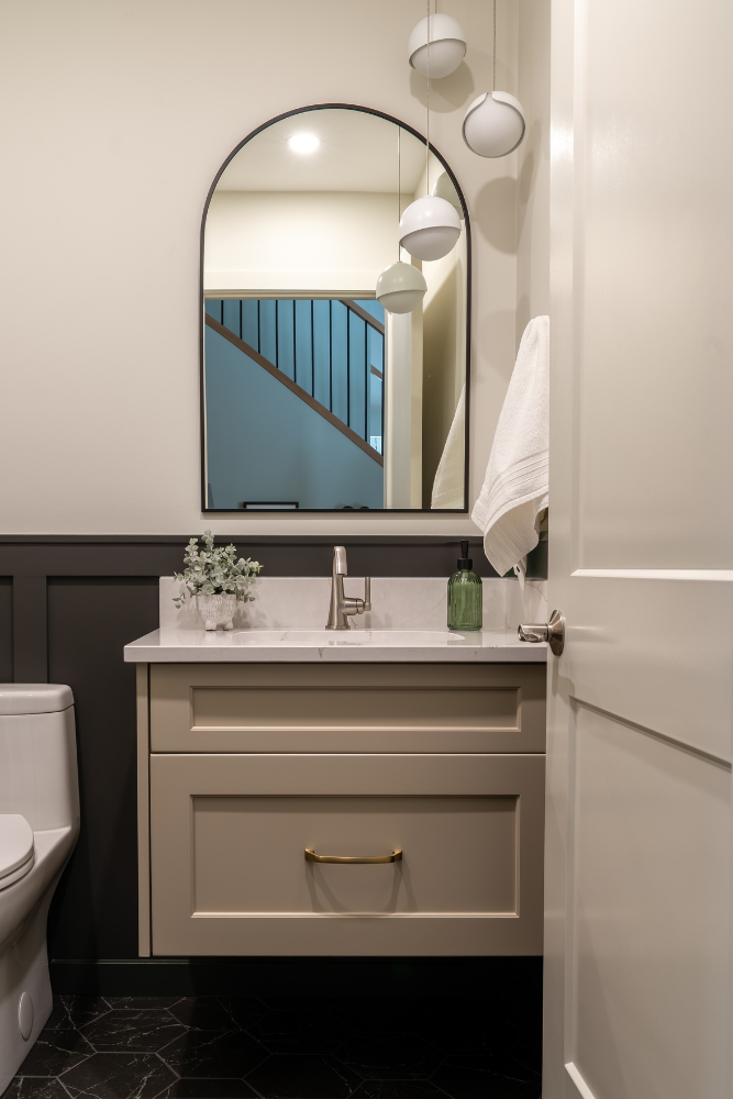 A close-up of a bathroom vanity with light beige cabinets, gold hardware, a black arched mirror reflecting a staircase, and modern multi-bulb pendant lighting.