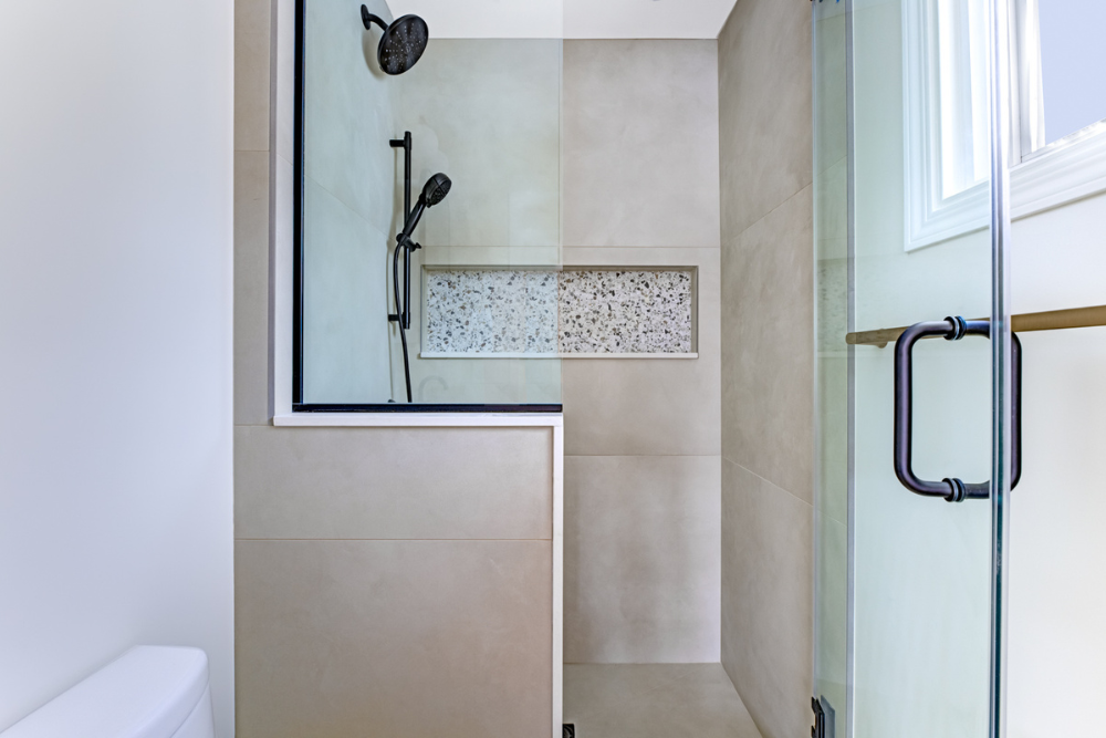 Close-up detail shot of a glass shower enclosure with large square taupe-colored wall tiles. It features matte black rainfall and hand-held shower heads, and a distinct multi-colored stone pebble mosaic tile soap niche. 