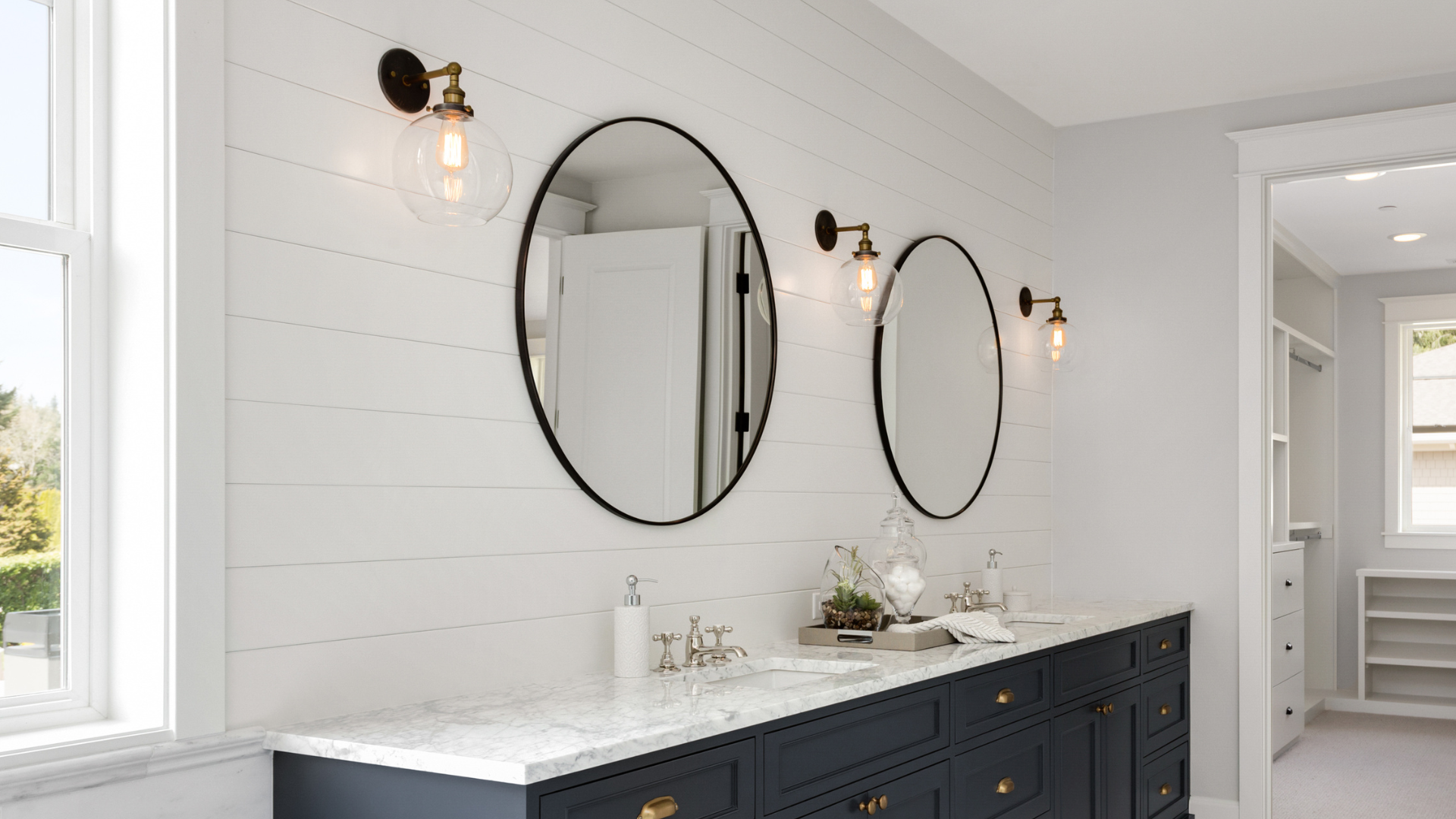 Modern bathroom with a double vanity featuring navy cabinets, marble countertops, round black-framed mirrors, shiplap wall, and brass globe sconces, showcasing a bright and elegant interior design.