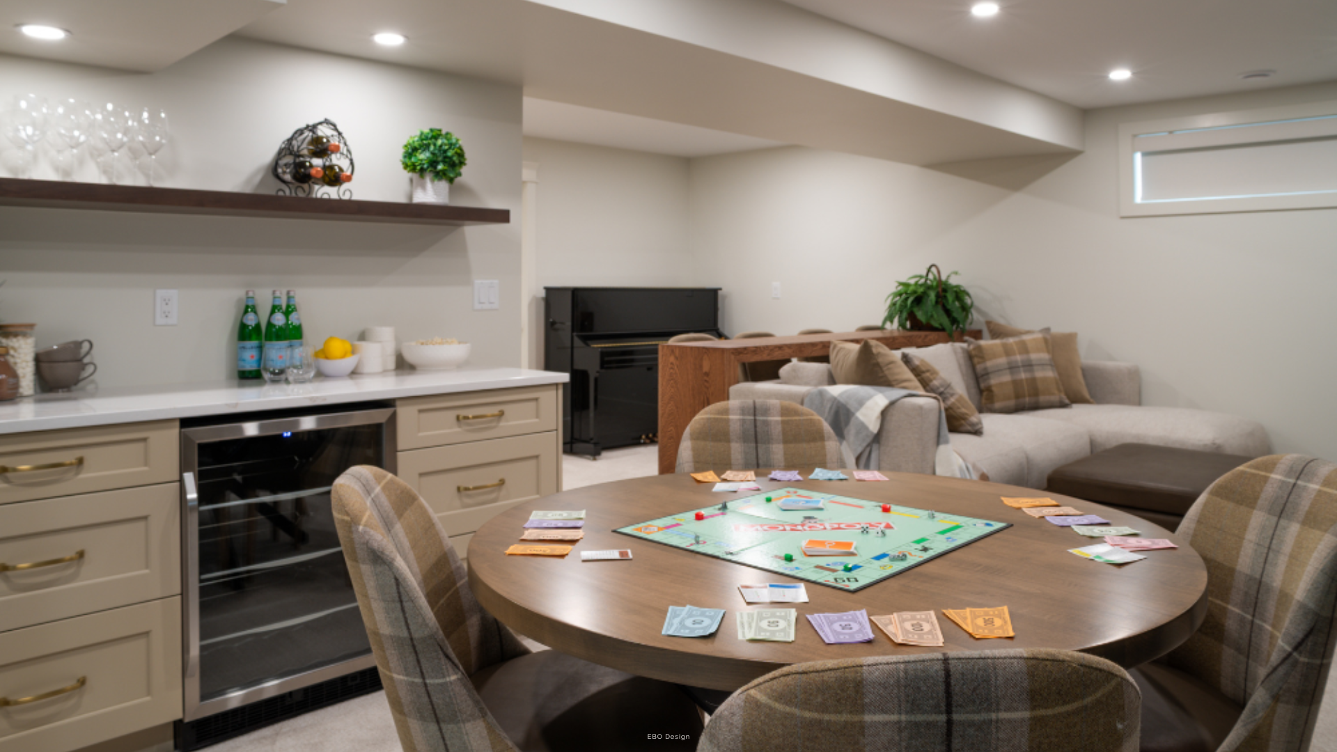 A modern finished basement featuring a round wooden table set for Monopoly, a built-in wet bar with a wine fridge, and a lounge area with a sofa and piano in the background