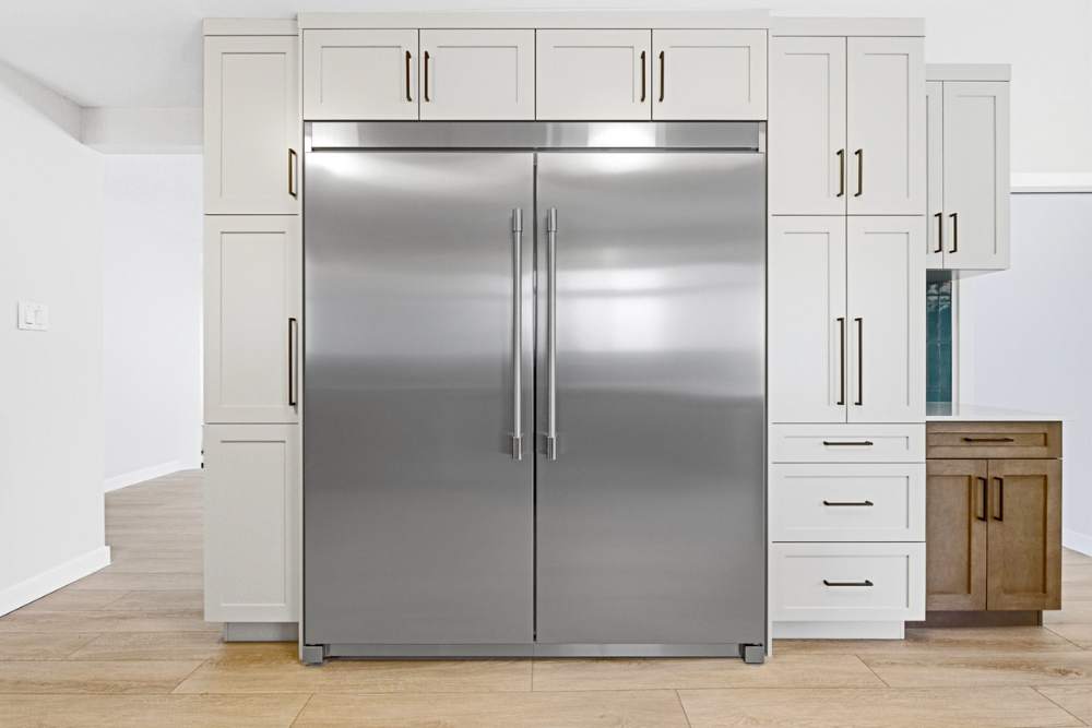A close-up frontal view of a custom-built stainless steel French door refrigerator with sleek horizontal handles, seamlessly integrated between floor-to-ceiling light grey and medium-toned wood cabinets. 
