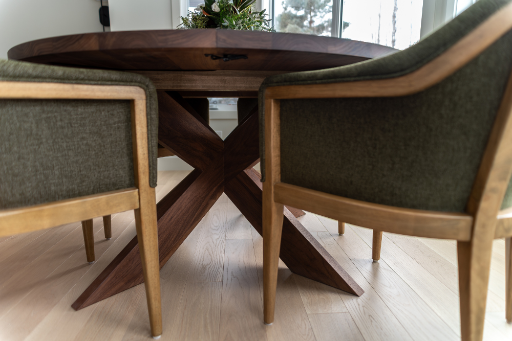 A low-angle shot showing the dark wood crisscross base structure of a round dining table, framed by two modern green upholstered chairs.