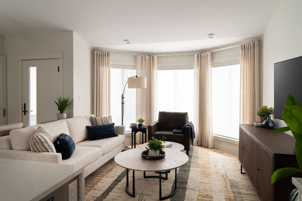 A cozy living room featuring a beige sectional sofa, nested marble-top coffee tables, and a dark leather armchair positioned by a sunlit bay window.
