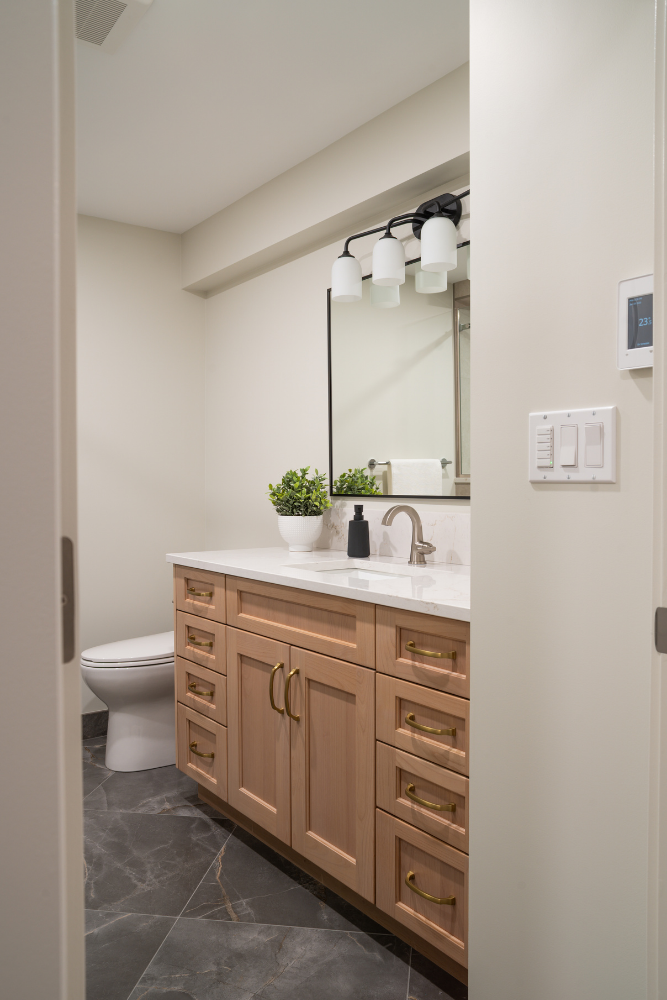 A view through a doorway into a modern bathroom featuring a light wood vanity, white countertop, dark grey marble floors, and a three-light globe fixture above the mirror.