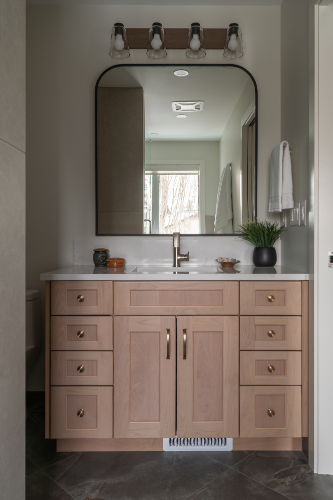 A straight-on shot of a custom light wood bathroom vanity with a white quartz countertop and single sink. It features elegant brass hardware, an arched black-framed mirror, and a multi-bulb light fixture.