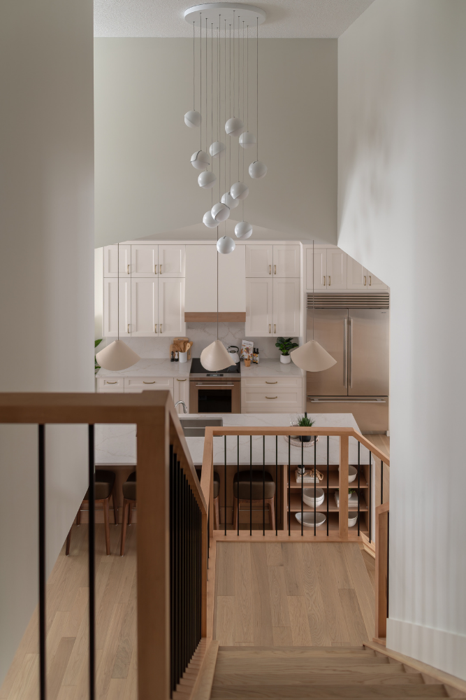 An elevated view looking down a modern open staircase with light wood steps and black railings. High ceilings are visible, featuring a large, artistic multi-globe pendant light fixture hanging above the first-floor kitchen.