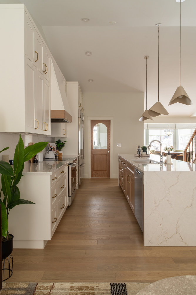 A wide perspective of an open-concept kitchen with white cabinets, wood accents on the island and range hood, unique pendant lights, and the pantry door in the background.