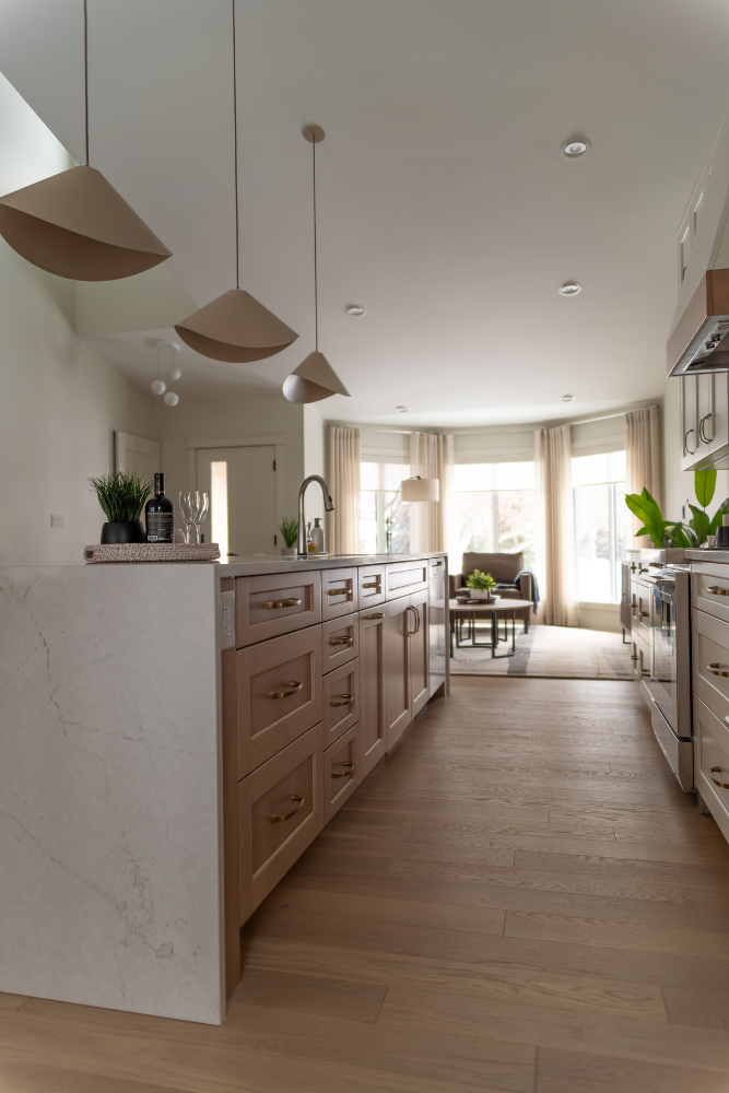 A view across a waterfall-edge kitchen island with modern pendant lights, showing the transition into a brightly lit living room with large bay windows.