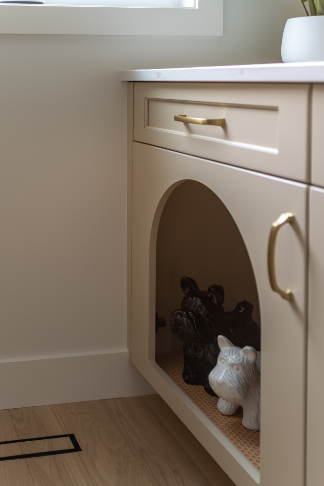 A playful detail of a light beige kitchen cabinet with brass hardware, featuring an arched floor-level cut-out cubby where two ceramic Scottie dog figurines are resting.