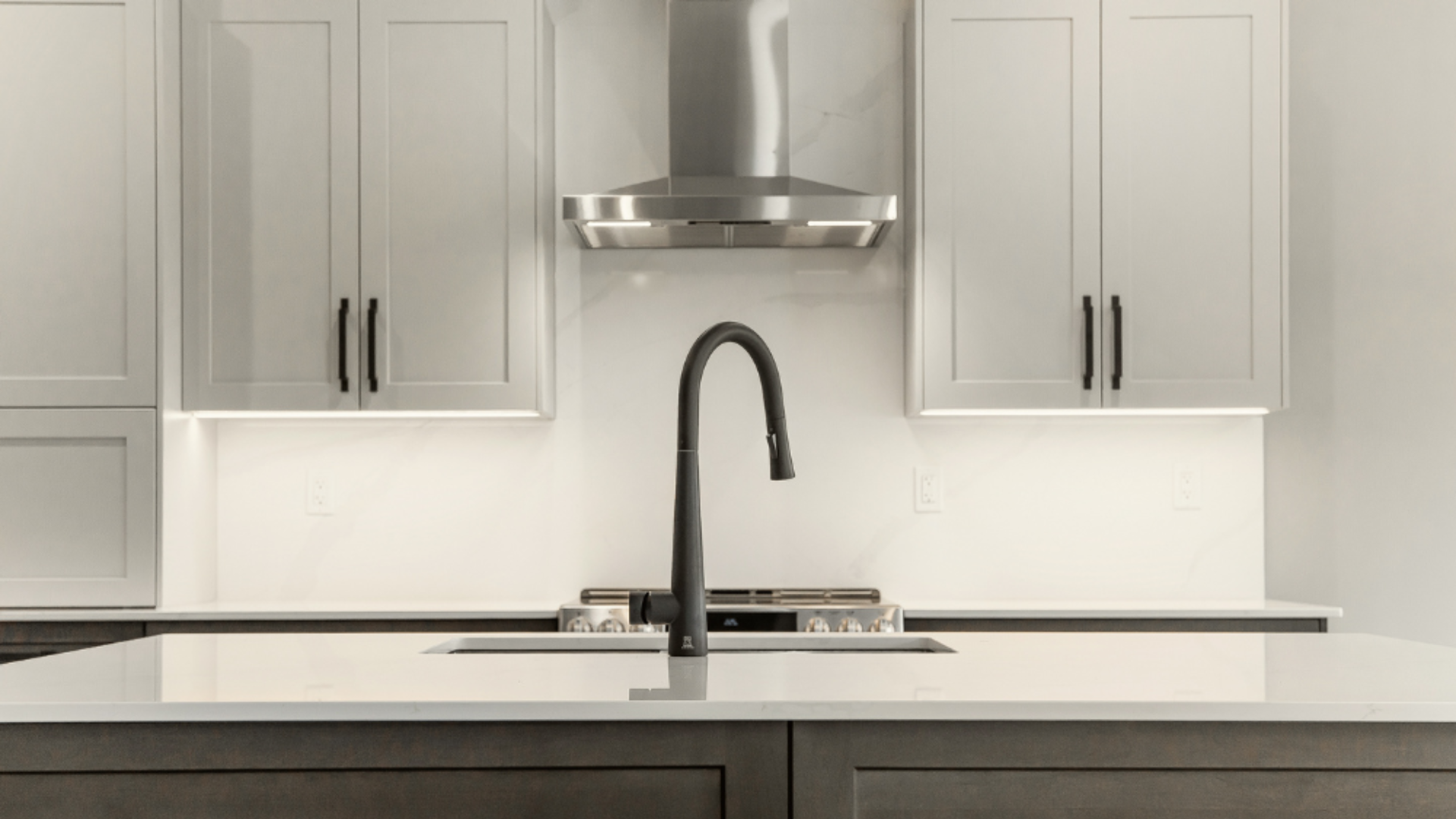 Close-up of a sleek matte black gooseneck faucet on a white quartz island countertop in front of a stainless steel range hood.