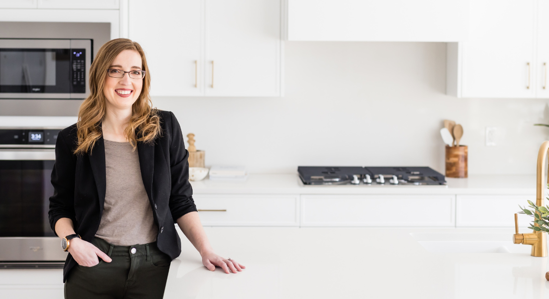 Chelsey Corea, Edmonton interior designer and founder of EBO Design, standing in a modern white kitchen with gold accents and custom cabinetry.