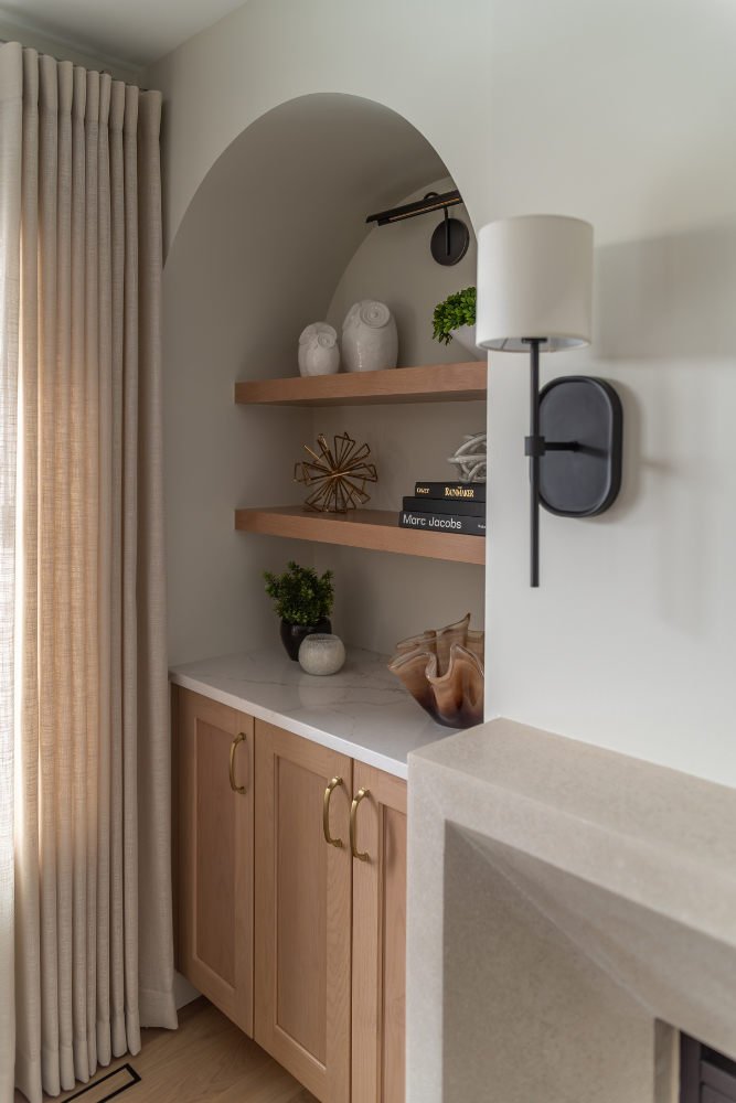 A detailed view of an arched built-in unit with light wood cabinets, quartz countertops, and floating wood shelves displaying various decorative items next to a black sconce.