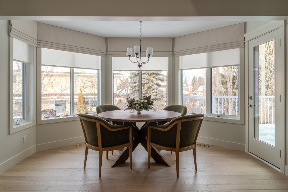 A bright breakfast nook set in a bay window with the shades open, featuring a round walnut-base table, green chairs, and a view of a wintery backyard.