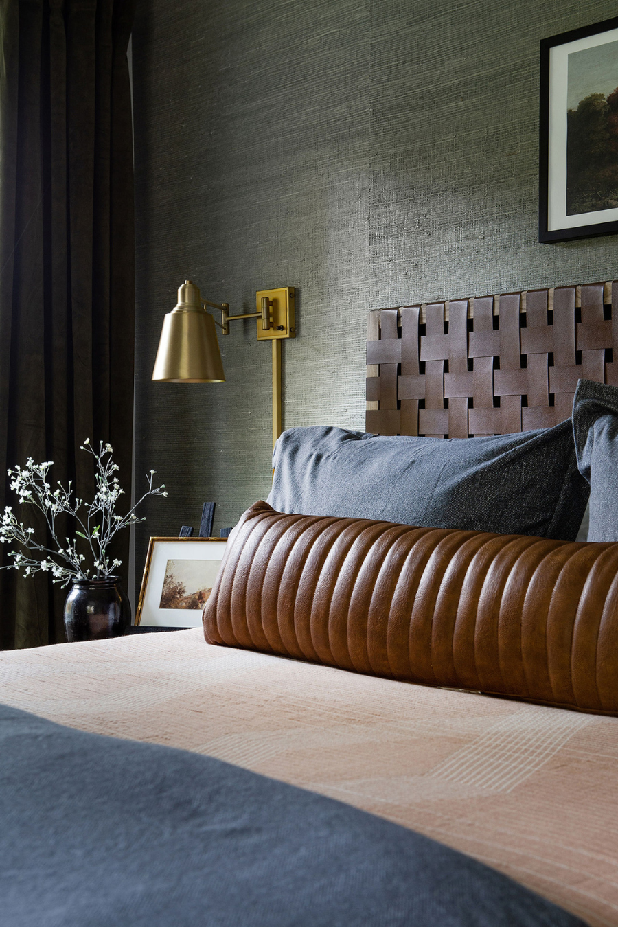 Modern cozy bedroom with a woven leather headboard, brass wall sconce, brown leather bolster pillow, textured bedding, and styled nightstand décor for a warm, sophisticated interior design look.