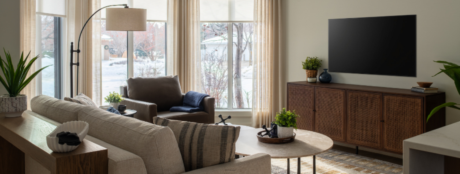 A bright, stylish modern living room with a textured sofa, a leather armchair, large floor-to-ceiling windows, and a dark wood media console.