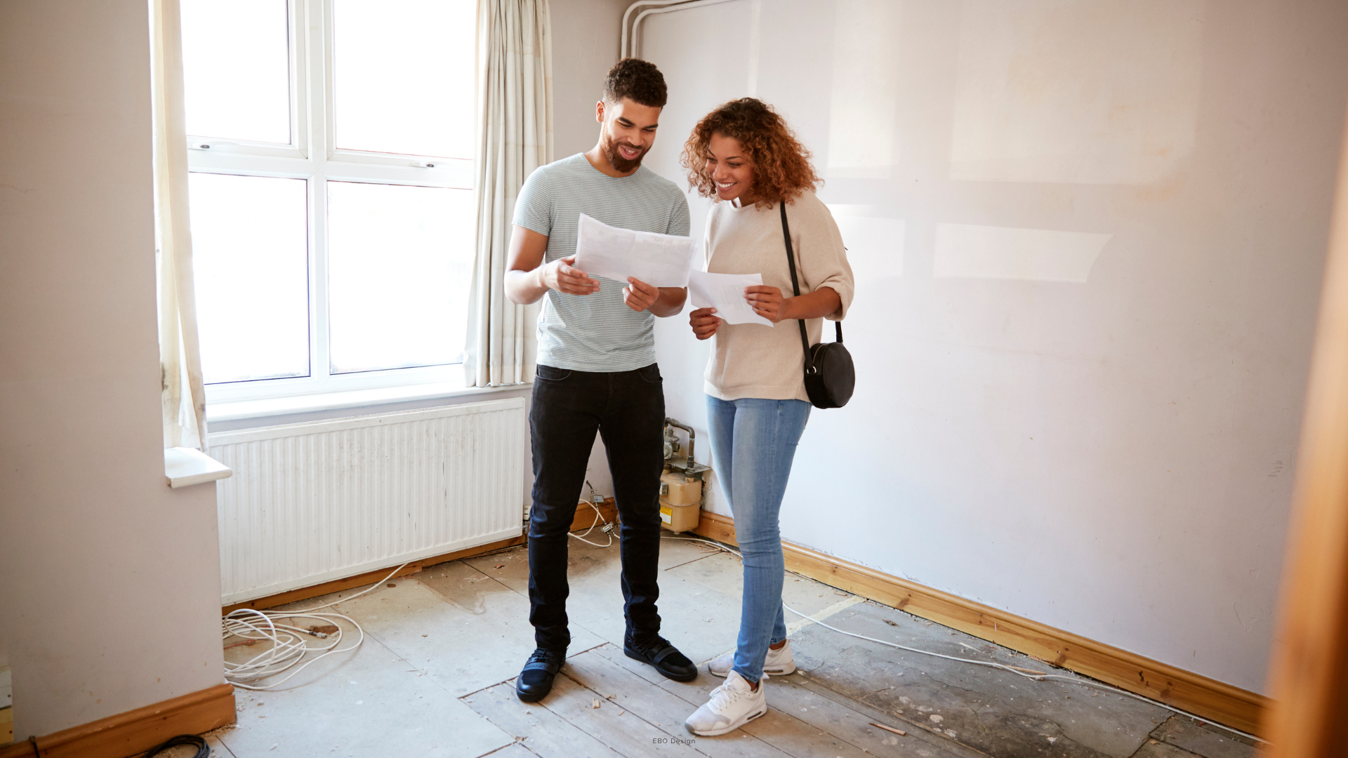 A happy young couple in an unfinished room, smiling as they look at floor plans together.