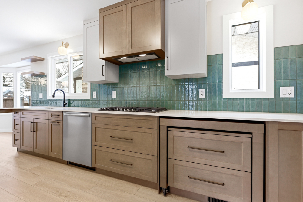A detailed perspective shot along a long kitchen wall, showcasing the combination of grey and medium-toned wood cabinetry with modern bar pulls. A matching wood range hood is integrated above a gas cooktop, with a teal tile backsplash and a stainless