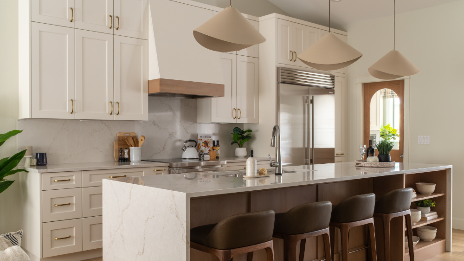 A bright modern transitional kitchen featuring white shaker cabinets, gold hardware, a stone waterfall-edge island with bar stools, and unique pendant lights.