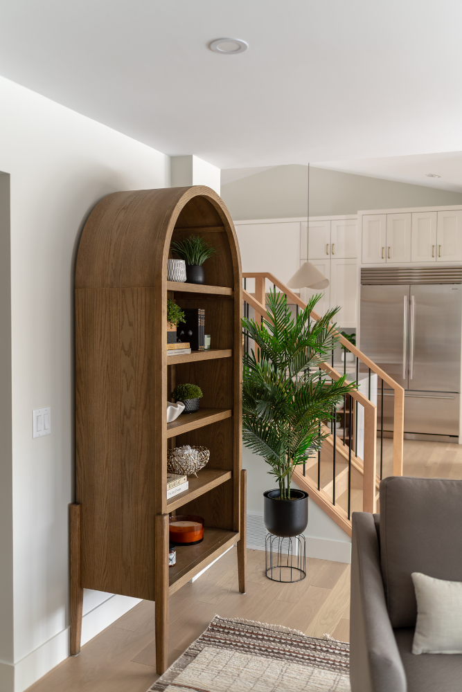 A shot of a large, free-standing, arched wood bookcase positioned next to a tall, potted artificial palm tree, with a modern staircase visible in the background.