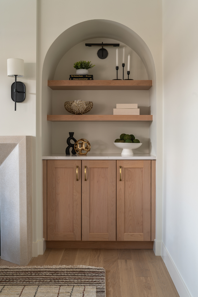 A more detailed view of an arched built-in with light wood floating shelves and lower cabinets, decorated with books, a gold bowl, candles, and a white dish filled with green decor.