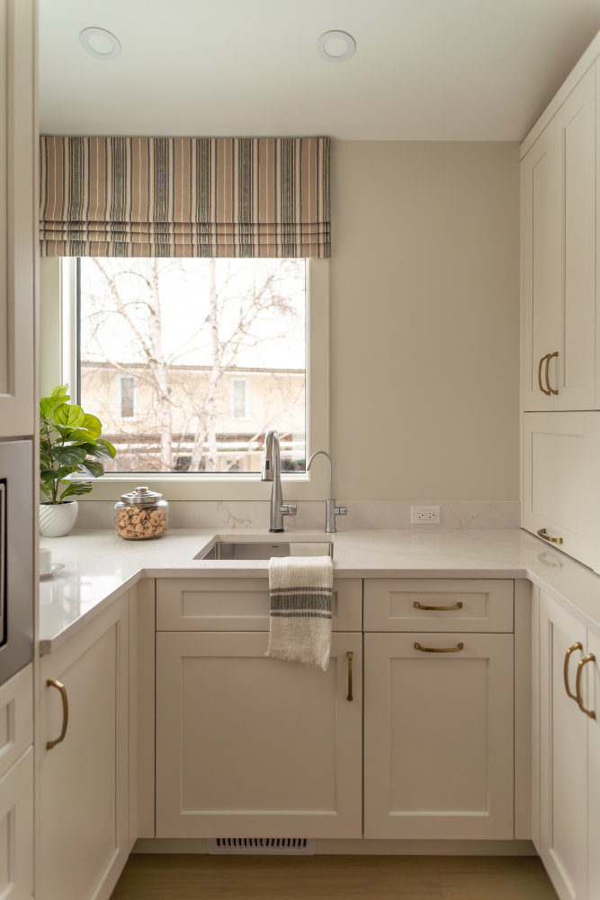 A shot of a kitchen sink area by a window, featuring a striped Roman shade, a faucet, a clear glass cookie jar full of cookies, and a potted plant.