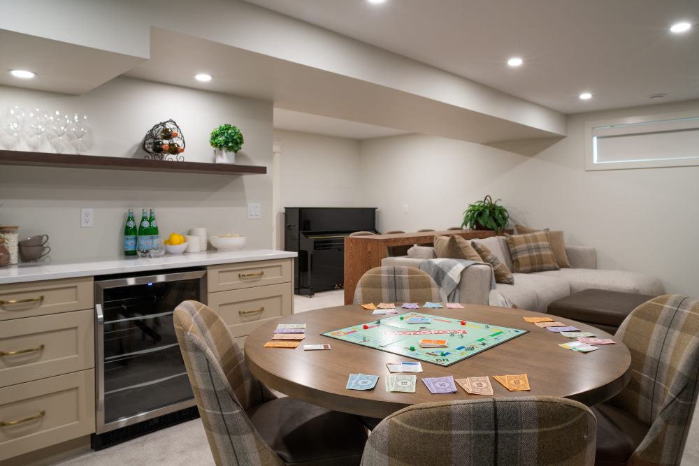 An inviting modern basement with a detailed wet bar, beverage cooler, and cabinetry next to a round game table with a Monopoly board set up. A sectional couch and piano are visible in the background.