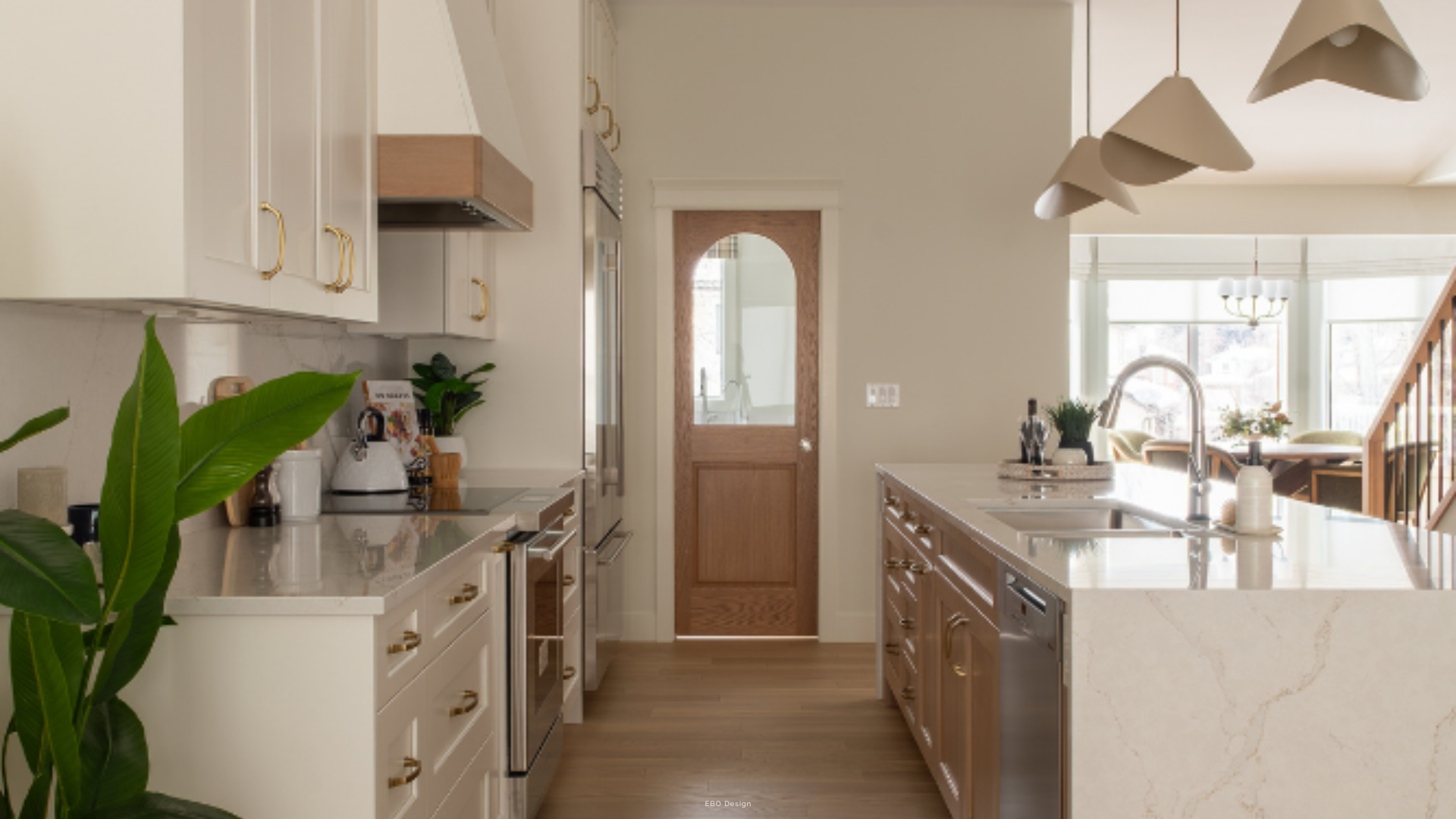 A modern kitchen featuring white shaker cabinets with gold hardware, a marble-patterned waterfall island with an undermount sink, and a view leading to a custom arched wooden door.