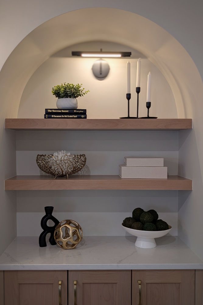 A stylized arched built-in unit with light wood shelves displaying books, plants, geometric ornaments, and candlesticks above a marble-top cabinet.