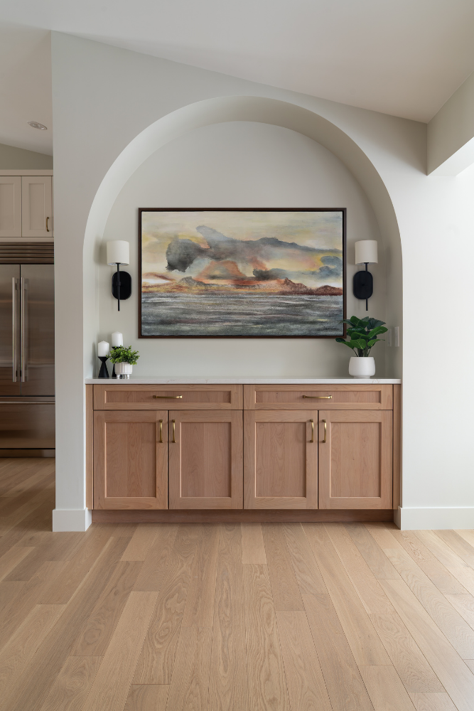A decorative arched built-in unit with light wood cabinets, a quartz counter, and a colorful abstract landscape painting centered between two black wall sconces.