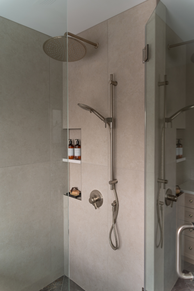 A detail shot within a custom shower enclosure, highlighting the brushed nickel plumbing fixtures, which include both a large overhead rainfall showerhead and an adjustable handheld shower wand with a slide bar.