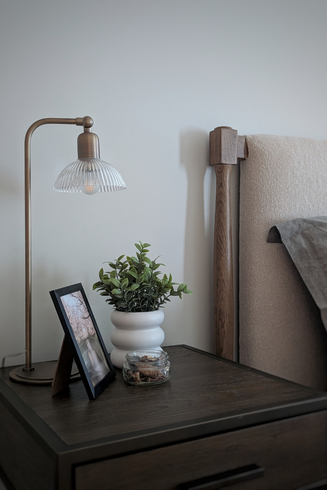 A close-up of a dark wood nightstand featuring a brass ribbed glass lamp, a framed picture, and a white pot with a faux plant next to a textured headboard.
