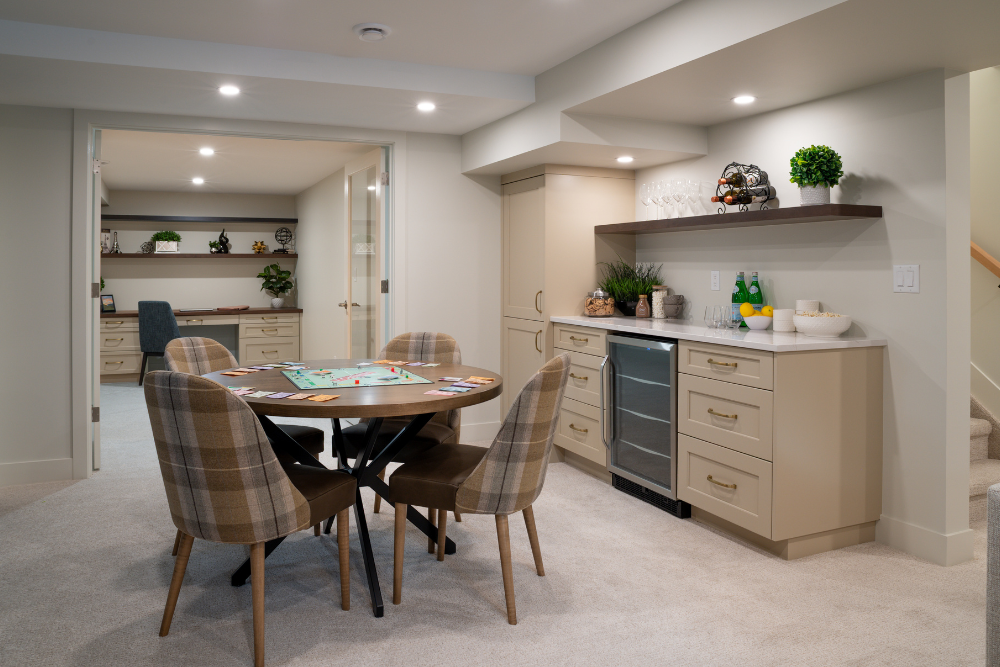 A view of a finished basement featuring a round wooden game table with plaid chairs, next to a built-in wet bar with a beverage cooler and cabinetry.