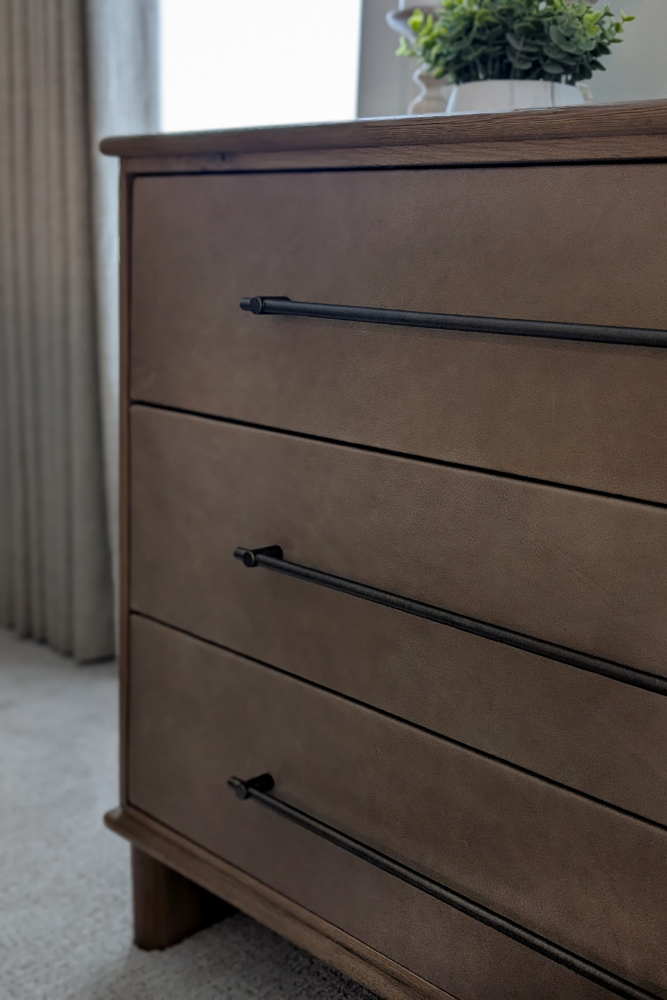A detail shot of a three-drawer dresser featuring rich brown leather drawer fronts and slim black horizontal bar handles.
