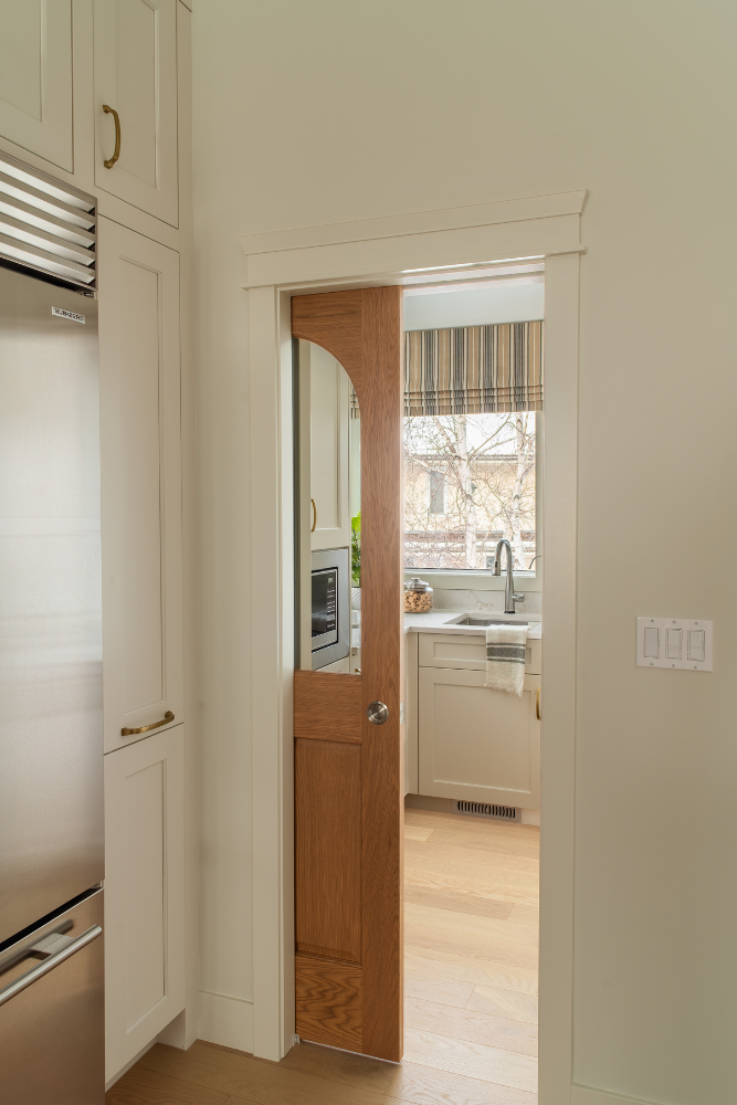 A view through an open arched wooden pantry door shows a kitchen sink, a striped window treatment, and bare winter trees outside.