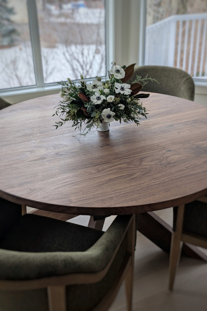 A detailed close-up of a round walnut wood dining table featuring a beautiful flower arrangement of white anemones and greenery.