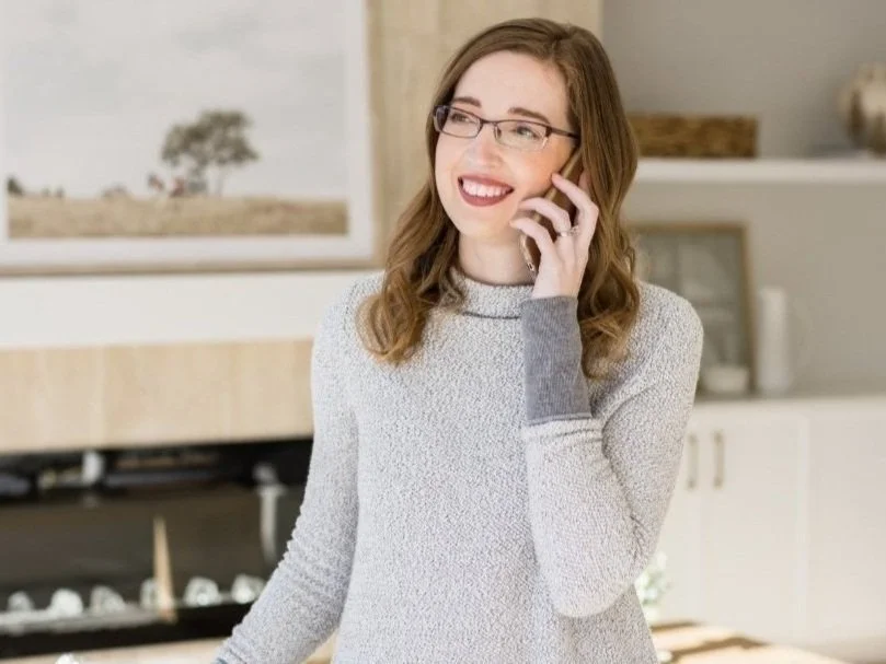 Interior designer speaking on the phone in a bright modern living room, standing in front of built-in shelving and neutral home decor elements.