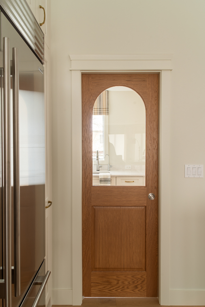 An arched wooden pantry door with glass panels sits next to a large stainless steel refrigerator in a modern kitchen with cream cabinets.