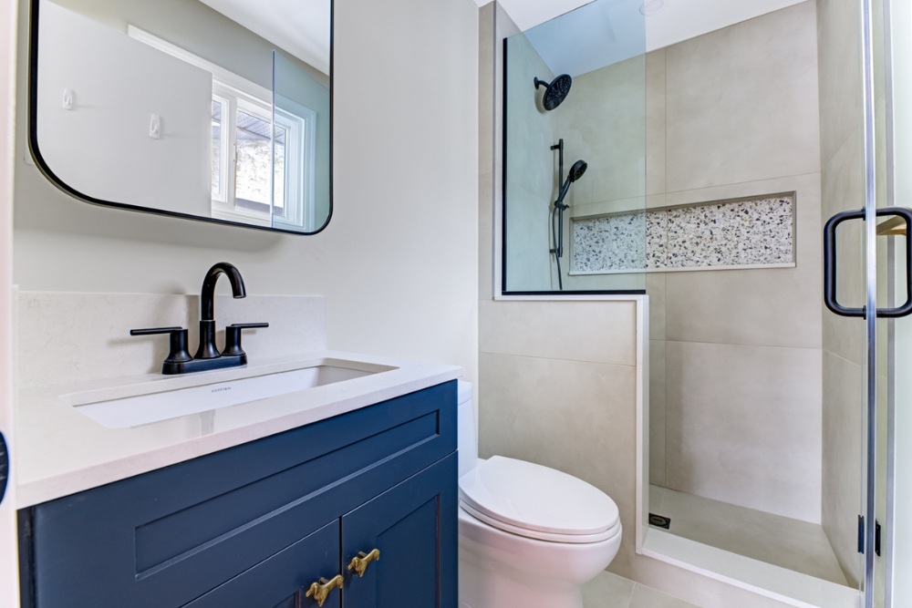 A modern bathroom interior with a floating navy blue vanity, a black faucet, a large rectangular black-framed mirror, a white toilet, and a glass-enclosed shower with large format taupe wall tiles, black fixtures, and a textured stone mosaic tile nic