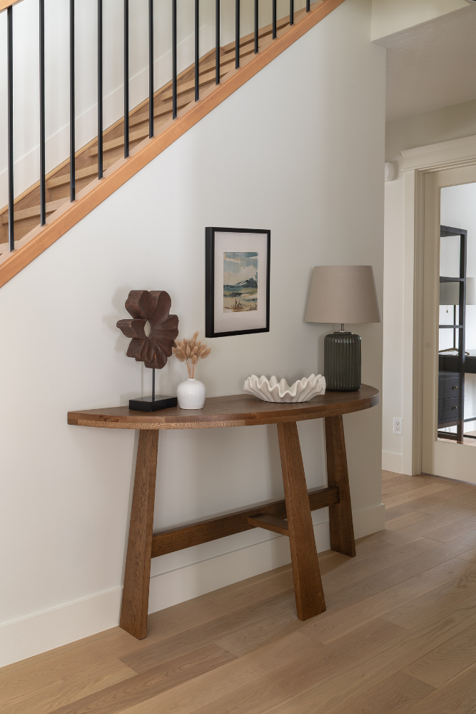 A mid-century modern wooden console table with angled legs against a wall, styled with pampas grass, a framed print, a textured lamp, and a fluted bowl near a staircase.